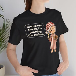Op de afbeelding: Een zwart t-shirt met een afbeelding van een stripfiguur in een rood-wit gestreepte pyjama en bijpassende slaapmuts. Het figuur houdt een brandende kaars en een bord koekjes vast, met de tekstballon: Everyone's asleep. I'm guarding the cookies.