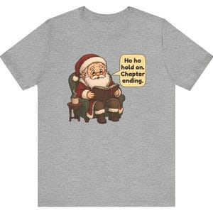 Op de afbeelding: Een grijs gem&ecirc;leerd t-shirt met een afbeelding van de Kerstman die in een groene fauteuil zit en een boek leest, naast een bijzettafeltje met een mok. Een tekstballon ernaast bevat de tekst: Ho ho hold on. Chapter ending.