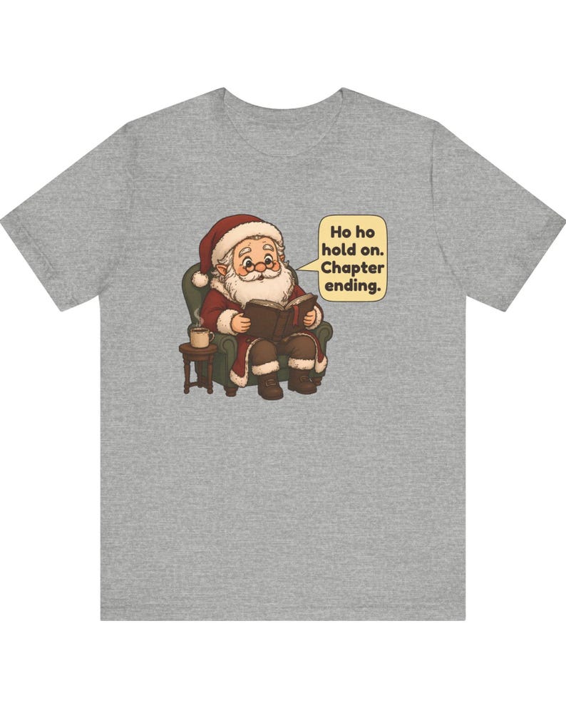Op de afbeelding: Een grijs gem&ecirc;leerd t-shirt met een afbeelding van de Kerstman die in een groene fauteuil zit en een boek leest, naast een bijzettafeltje met een mok. Een tekstballon ernaast bevat de tekst: Ho ho hold on. Chapter ending.