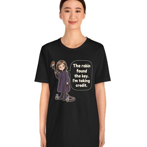May include: A black t-shirt featuring a graphic illustration of a person in a dark coat holding a large key with a robin perched on it. A speech bubble contains the text: The robin found the key. I'm taking credit. The design includes small flowers.