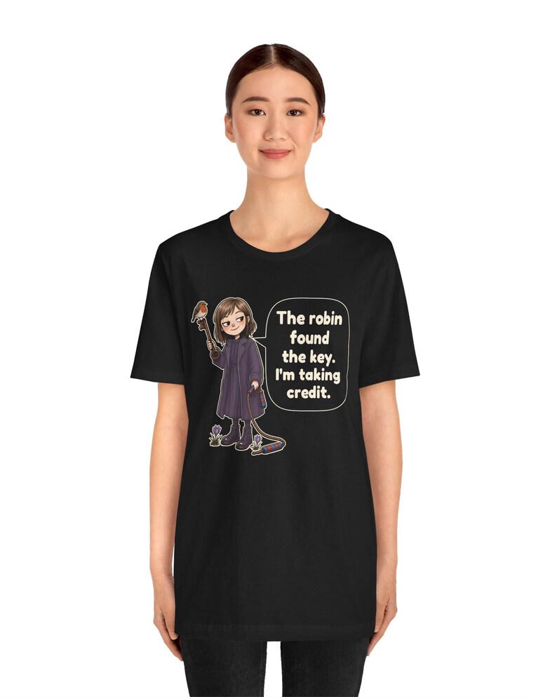 May include: A black t-shirt featuring a graphic illustration of a person in a dark coat holding a large key with a robin perched on it. A speech bubble contains the text: The robin found the key. I'm taking credit. The design includes small flowers.