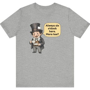 May include: A heather gray t-shirt featuring a cartoon character wearing a top hat, brown vest, and checkered pants, holding a teacup. A speech bubble next to the character contains the text: Always six o'clock here. More tea?