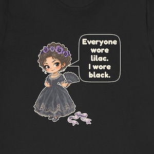 May include: A black t-shirt featuring a cartoon illustration of a character in a dark dress with a floral headpiece and a hand fan. A speech bubble contains the text: Everyone wore lilac. I wore black. A small purple ribbon bow is below the figure.