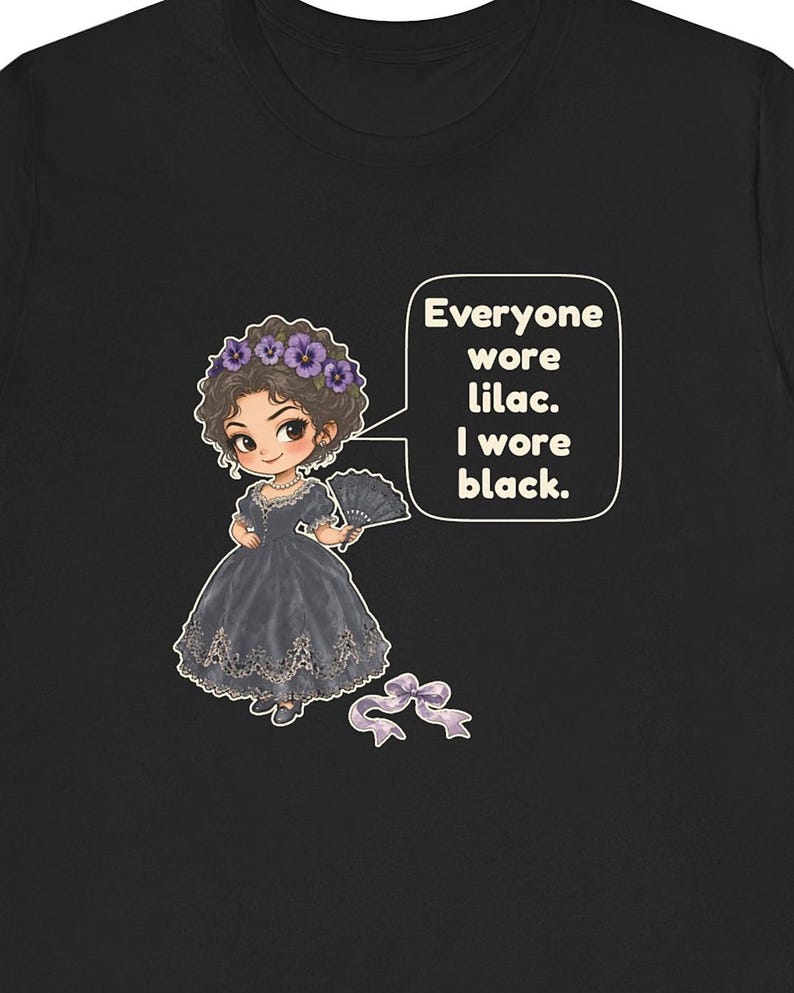 May include: A black t-shirt featuring a cartoon illustration of a character in a dark dress with a floral headpiece and a hand fan. A speech bubble contains the text: Everyone wore lilac. I wore black. A small purple ribbon bow is below the figure.