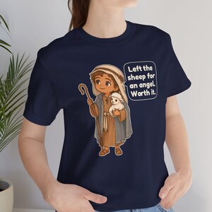 May include: A navy blue t-shirt featuring a cartoon illustration of a shepherd holding a small white lamb and a wooden staff. The shepherd wears a brown tunic and a head covering. A speech bubble contains the text: Left the sheep for an angel. Worth it.