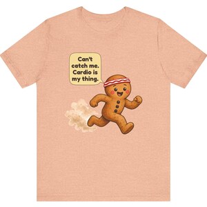 May include: A peach-coloured t-shirt featuring a graphic of a running gingerbread person wearing a red and white headband. A speech bubble above the character contains the text: Can't catch me. Cardio is my thing. A small dust cloud is visible behind the figure.