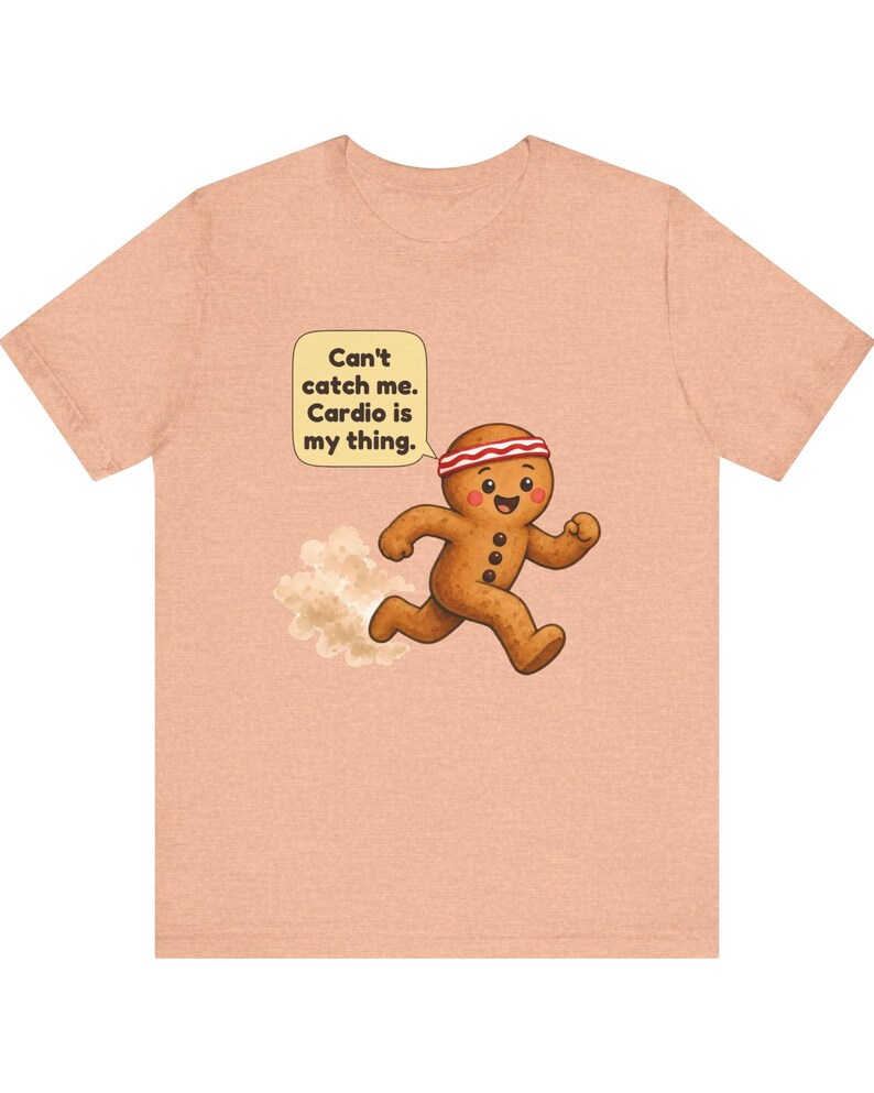 May include: A peach-coloured t-shirt featuring a graphic of a running gingerbread person wearing a red and white headband. A speech bubble above the character contains the text: Can't catch me. Cardio is my thing. A small dust cloud is visible behind the figure.