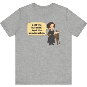 May include: A heather gray t-shirt featuring a cartoon illustration of a person in a black dress standing next to an easel with a painting. A speech bubble contains the text: Left the husband. Kept the paintbrushes.