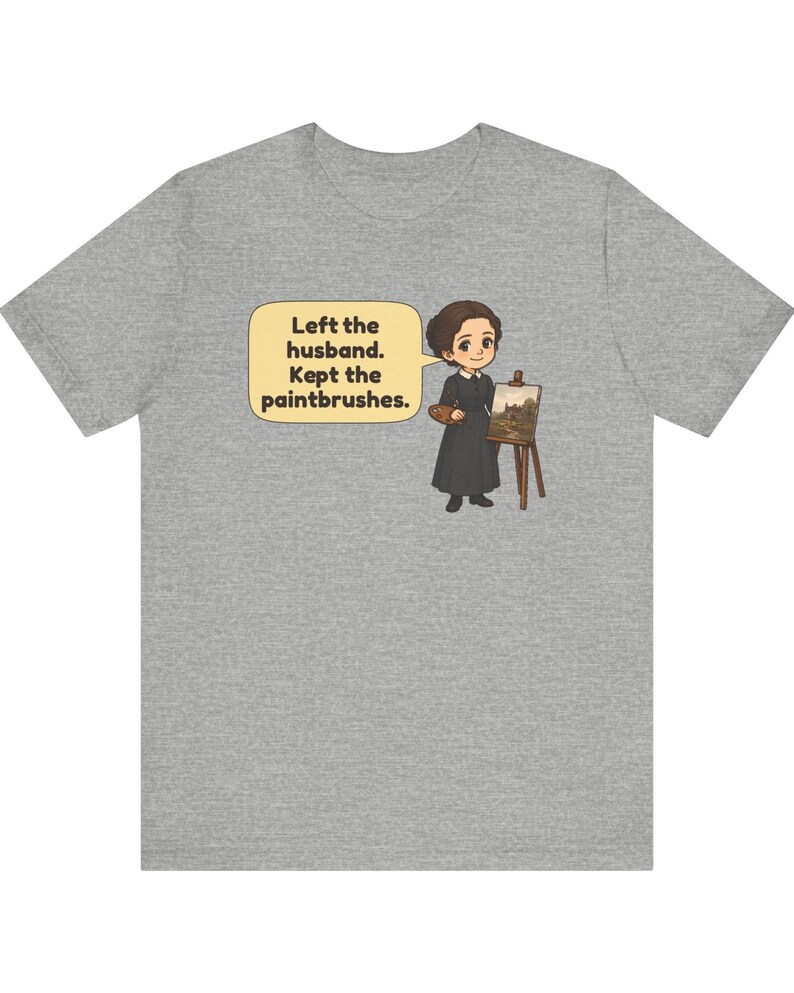 May include: A heather gray t-shirt featuring a cartoon illustration of a person in a black dress standing next to an easel with a painting. A speech bubble contains the text: Left the husband. Kept the paintbrushes.