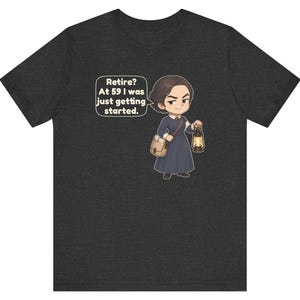 May include: A dark heather gray t-shirt featuring a cartoon illustration of a person in a long dark dress carrying a satchel and a lit lantern. A speech bubble next to the character contains the text: Retire? At 59 I was just getting started.