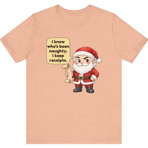 May include: A peach-colored t-shirt featuring a cartoon Santa Claus holding a scroll. A speech bubble next to Santa contains the text: I know who's been naughty. I keep receipts. The design is centered on the front of the short-sleeved crew neck shirt.