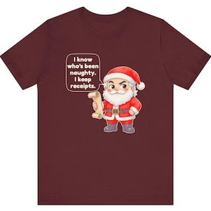 May include: A maroon short-sleeve t-shirt featuring a cartoon Santa Claus holding a scroll. A speech bubble above Santa contains the text: I know who's been naughty. I keep receipts. The graphic is centered on the front of the shirt.