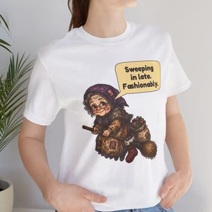 Puede incluir: Una camiseta blanca con una ilustraci&oacute;n gr&aacute;fica de una persona mayor con pa&ntilde;uelo morado y gafas, montada en una escoba. Un bocadillo amarillo sobre el personaje contiene el texto en negro: Sweeping in late. Fashionably.