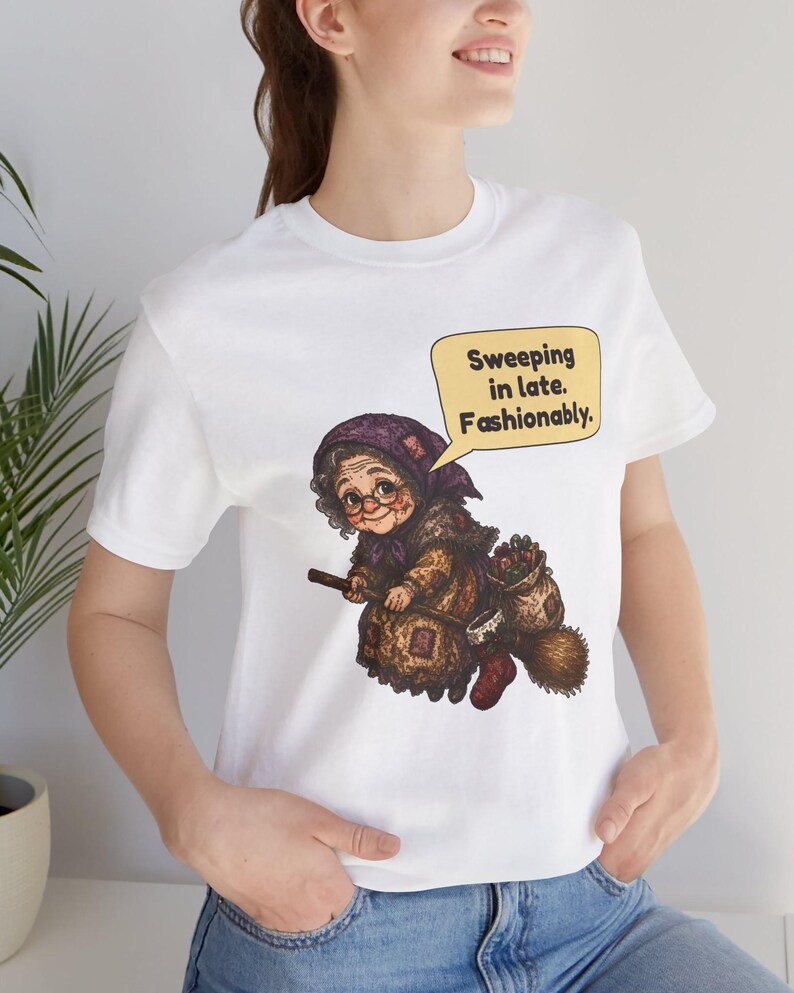 Puede incluir: Una camiseta blanca con una ilustraci&oacute;n gr&aacute;fica de una persona mayor con pa&ntilde;uelo morado y gafas, montada en una escoba. Un bocadillo amarillo sobre el personaje contiene el texto en negro: Sweeping in late. Fashionably.