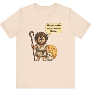 May include: A light beige t-shirt featuring a graphic of a one-eyed cyclops holding a wooden staff and standing next to a small white sheep and a large round piece of cheese. A speech bubble above the cyclops contains the text: Guests ate my cheese. Rude.
