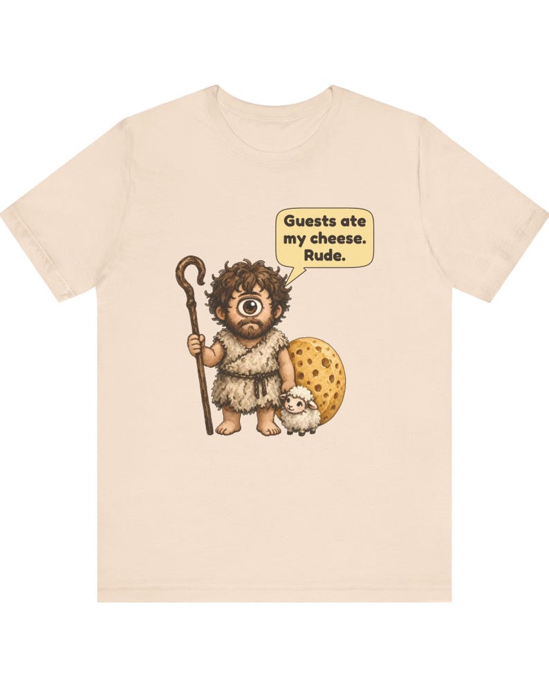 May include: A light beige t-shirt featuring a graphic of a one-eyed cyclops holding a wooden staff and standing next to a small white sheep and a large round piece of cheese. A speech bubble above the cyclops contains the text: Guests ate my cheese. Rude.