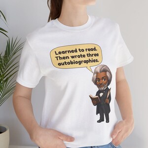 Puede incluir: Una camiseta blanca con una ilustraci&oacute;n de dibujos animados de Frederick Douglass sosteniendo un libro y una pluma. Un globo de texto sobre la figura contiene el texto: Learned to read. Then wrote three autobiographies.