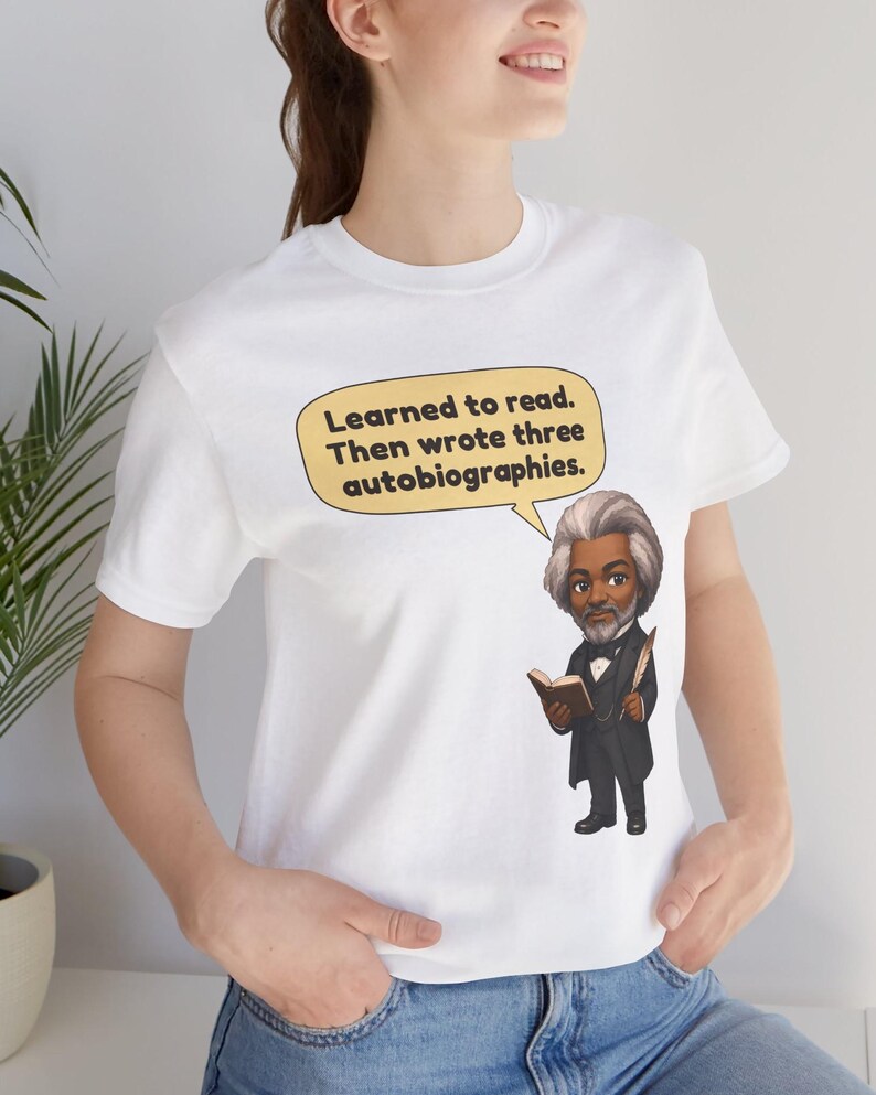 Puede incluir: Una camiseta blanca con una ilustraci&oacute;n de dibujos animados de Frederick Douglass sosteniendo un libro y una pluma. Un globo de texto sobre la figura contiene el texto: Learned to read. Then wrote three autobiographies.