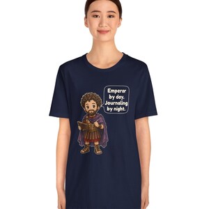 K&ouml;nnte beinhalten: Ein dunkelblaues Kurzarm-T-Shirt mit einer Cartoon-Illustration einer b&auml;rtigen Figur in r&ouml;mischer R&uuml;stung und violettem Umhang, die ein Tagebuch h&auml;lt. Eine Sprechblase enth&auml;lt den Text: Emperor by day. Journaling by night.