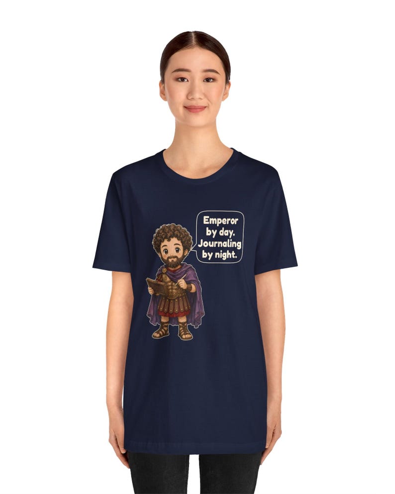 K&ouml;nnte beinhalten: Ein dunkelblaues Kurzarm-T-Shirt mit einer Cartoon-Illustration einer b&auml;rtigen Figur in r&ouml;mischer R&uuml;stung und violettem Umhang, die ein Tagebuch h&auml;lt. Eine Sprechblase enth&auml;lt den Text: Emperor by day. Journaling by night.