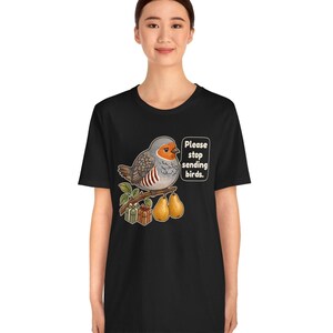 K&ouml;nnte beinhalten: Ein schwarzes Kurzarm-T-Shirt mit einer Grafik eines grauen und orangefarbenen Vogels, der auf einem Ast mit zwei Birnen und zwei kleinen Geschenkkartons sitzt. Eine Sprechblase neben dem Vogel enth&auml;lt den Text: "Please stop sending birds."