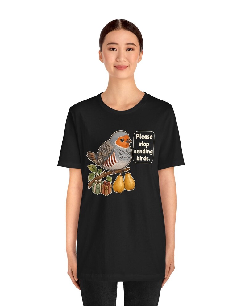 K&ouml;nnte beinhalten: Ein schwarzes Kurzarm-T-Shirt mit einer Grafik eines grauen und orangefarbenen Vogels, der auf einem Ast mit zwei Birnen und zwei kleinen Geschenkkartons sitzt. Eine Sprechblase neben dem Vogel enth&auml;lt den Text: "Please stop sending birds."
