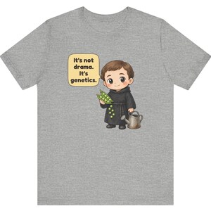 May include: A heather gray t-shirt featuring a cartoon illustration of a monk holding a pea pod and a watering can. A speech bubble next to the character contains the text: It's not drama. It's genetics. The design is centered on the front.