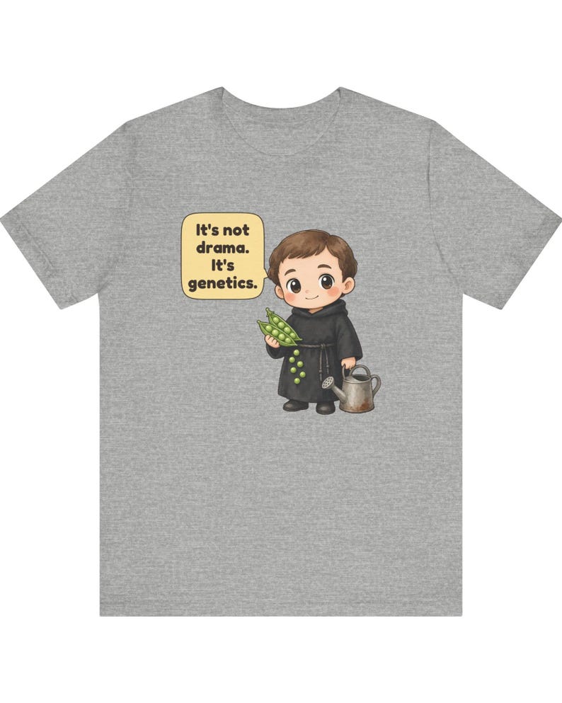 May include: A heather gray t-shirt featuring a cartoon illustration of a monk holding a pea pod and a watering can. A speech bubble next to the character contains the text: It's not drama. It's genetics. The design is centered on the front.