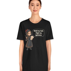 May include: A black short-sleeved t-shirt featuring a cartoon illustration of a person with a braid, wearing a brown dress and apron, holding a paper. A speech bubble contains the text: Sold my hair. Kept my opinions.