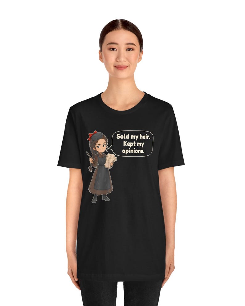 May include: A black short-sleeved t-shirt featuring a cartoon illustration of a person with a braid, wearing a brown dress and apron, holding a paper. A speech bubble contains the text: Sold my hair. Kept my opinions.