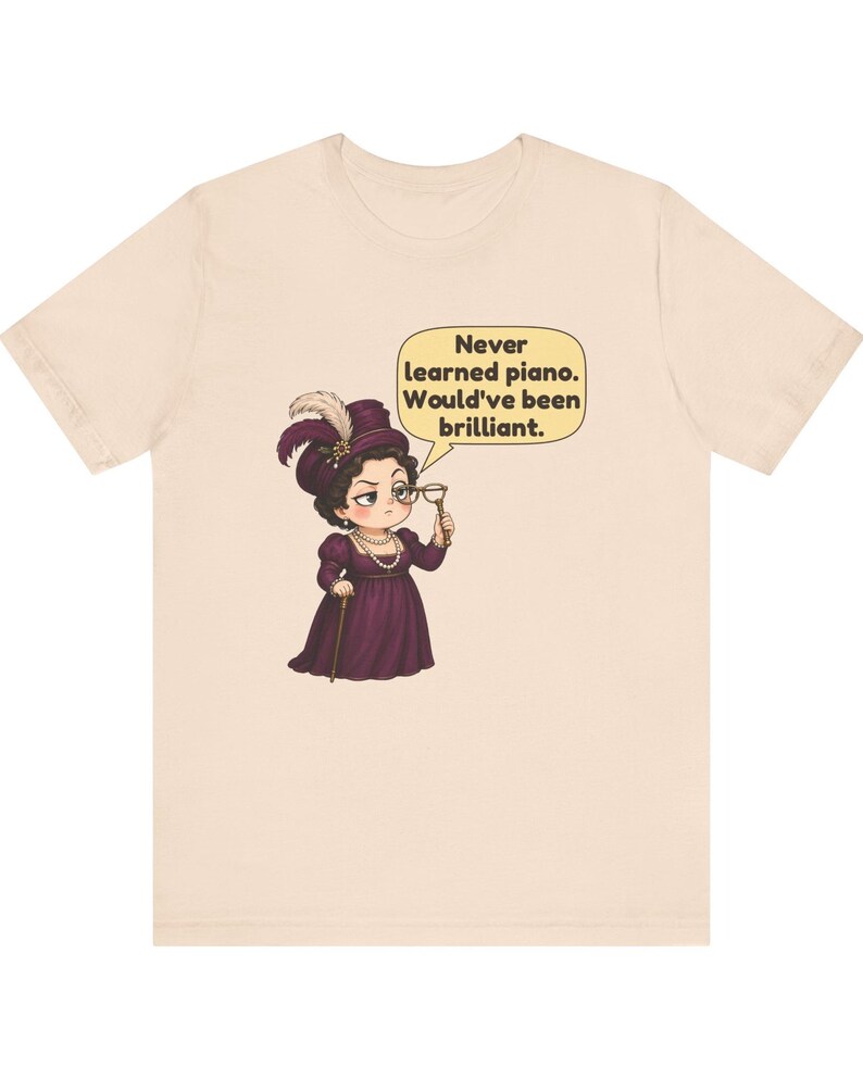 May include: A light beige t-shirt featuring a cartoon illustration of a person in a dark purple dress and hat, holding a monocle. A speech bubble contains the text: Never learned piano. Would've been brilliant.