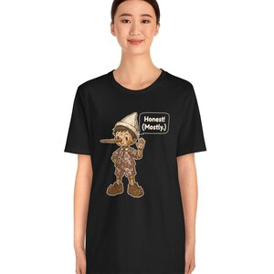 K&ouml;nnte beinhalten: Ein schwarzes Kurzarm-T-Shirt mit einer Grafik einer Holzpuppe mit langer Nase, die einen Hut und gemusterte Kleidung tr&auml;gt. Neben der Figur befindet sich eine wei&szlig;e Sprechblase mit dem Text "Honest! (Mostly.)"