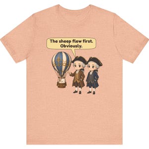 May include: A peach-colored t-shirt featuring a graphic of two historical figures in tricorn hats standing next to a hot air balloon with a sheep in the basket. A speech bubble above them contains the text: The sheep flew first. Obviously.