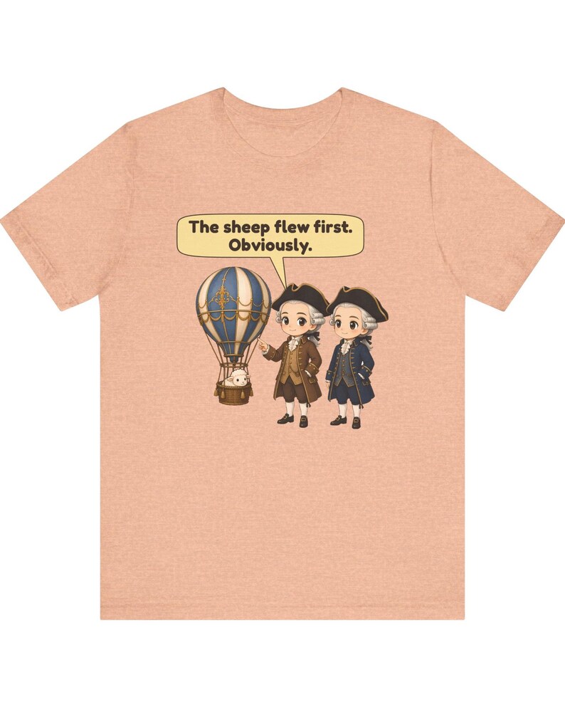 May include: A peach-colored t-shirt featuring a graphic of two historical figures in tricorn hats standing next to a hot air balloon with a sheep in the basket. A speech bubble above them contains the text: The sheep flew first. Obviously.
