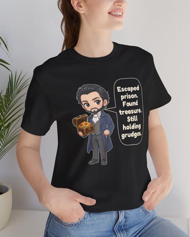 May include: A black t-shirt featuring a cartoon illustration of a bearded man in a blue coat holding a treasure chest. A speech bubble contains the text: Escaped prison. Found treasure. Still holding grudges. The design is centered on the chest.