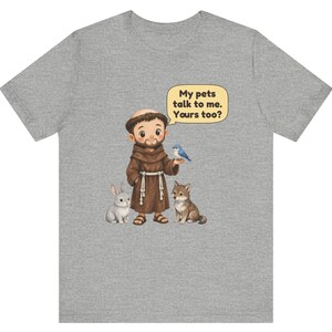 May include: A heather gray t-shirt featuring a cartoon illustration of a monk in a brown robe standing with a rabbit, a wolf, and a blue bird. A speech bubble above the monk contains the text: My pets talk to me. Yours too?