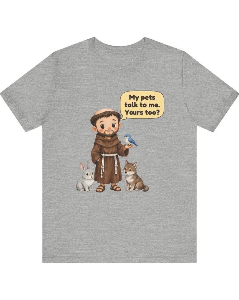May include: A heather gray t-shirt featuring a cartoon illustration of a monk in a brown robe standing with a rabbit, a wolf, and a blue bird. A speech bubble above the monk contains the text: My pets talk to me. Yours too?
