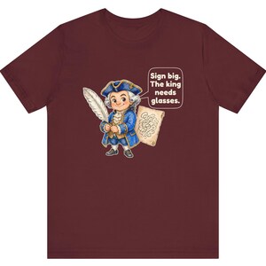 May include: A maroon short-sleeve t-shirt featuring a cartoon illustration of a person in an 18th-century blue coat and tricorn hat holding a quill. A speech bubble contains the text: Sign big. The king needs glasses.