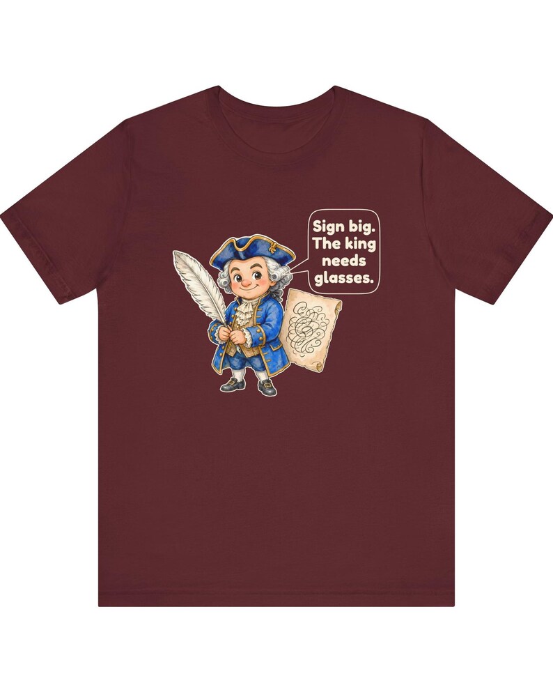 May include: A maroon short-sleeve t-shirt featuring a cartoon illustration of a person in an 18th-century blue coat and tricorn hat holding a quill. A speech bubble contains the text: Sign big. The king needs glasses.