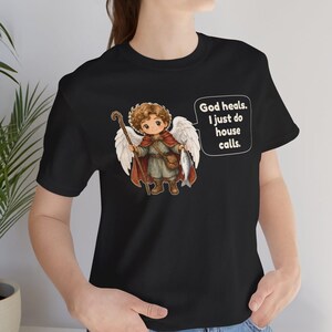May include: A black t-shirt featuring a graphic of a curly-haired angel with white wings, wearing a red cape and brown boots, holding a wooden staff and a fish. A speech bubble contains the text: God heals. I just do house calls.