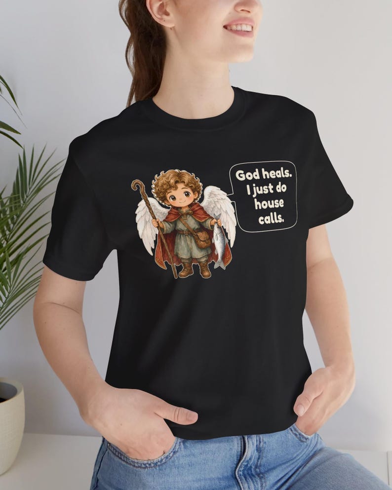 May include: A black t-shirt featuring a graphic of a curly-haired angel with white wings, wearing a red cape and brown boots, holding a wooden staff and a fish. A speech bubble contains the text: God heals. I just do house calls.