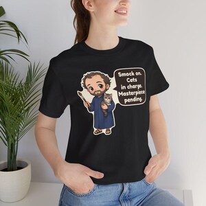 May include: A black short-sleeve t-shirt featuring a cartoon illustration of a bearded artist in a blue smock holding a paintbrush and a small cat. A speech bubble contains the text: Smock on. Cats in charge. Masterpiece pending.