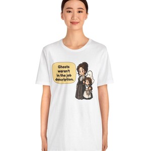 Op de afbeelding: Een wit t-shirt met korte mouwen en een afbeelding van een vrouw en een kind met een pop, vergezeld door een klein spookje. Een tekstballon bevat de tekst: Ghosts weren't in the job description. Het ontwerp staat centraal op de borst.