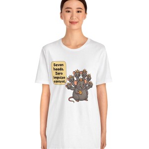 May include: A white t-shirt featuring a graphic of a gray seven-headed rat wearing small gold crowns and holding a piece of yellow cheese. A speech bubble next to the rat contains the black text: Seven heads. Zero impulse control.