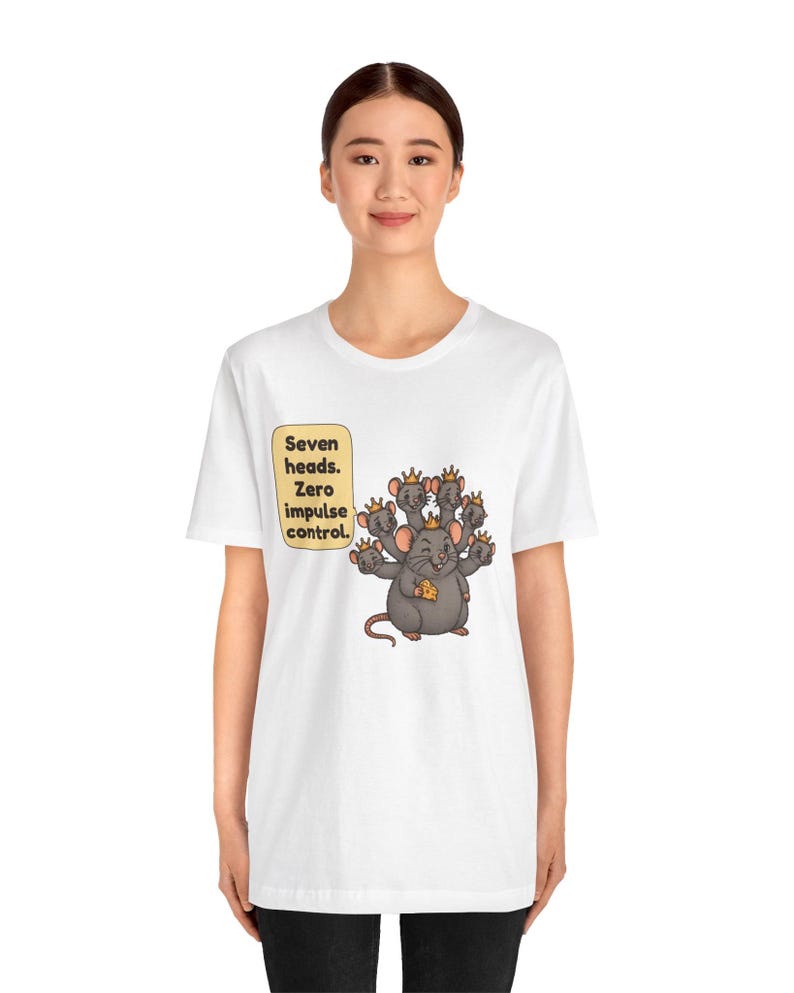 May include: A white t-shirt featuring a graphic of a gray seven-headed rat wearing small gold crowns and holding a piece of yellow cheese. A speech bubble next to the rat contains the black text: Seven heads. Zero impulse control.