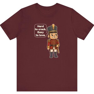 Pu&ograve; includere: Una t-shirt a maniche corte color bordeaux con la grafica di un soldatino schiaccianoci in uniforme rossa. Un fumetto accanto al personaggio contiene il testo: Hard to crack. Easy to love.