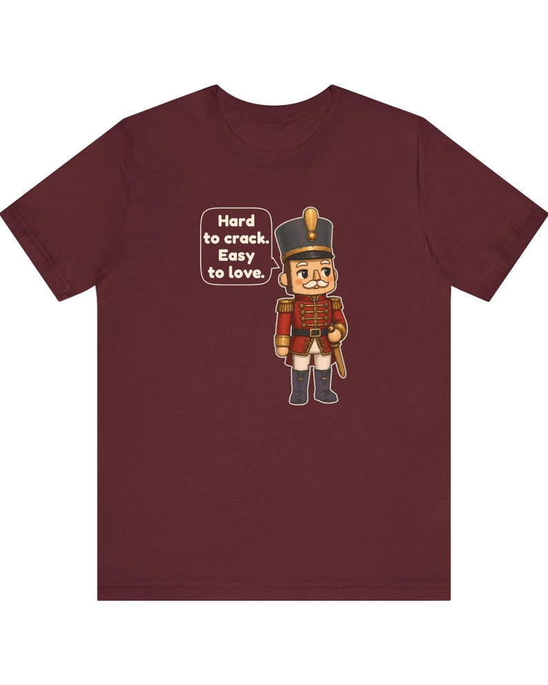 Pu&ograve; includere: Una t-shirt a maniche corte color bordeaux con la grafica di un soldatino schiaccianoci in uniforme rossa. Un fumetto accanto al personaggio contiene il testo: Hard to crack. Easy to love.