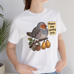 K&ouml;nnte beinhalten: Ein wei&szlig;es T-Shirt mit einer Grafik eines runden Vogels, der auf einem Ast mit zwei Birnen und zwei kleinen Geschenkkartons sitzt. Eine Sprechblase neben dem Vogel enth&auml;lt den Text: Please stop sending birds. Das Design nutzt Erdt&ouml;ne.