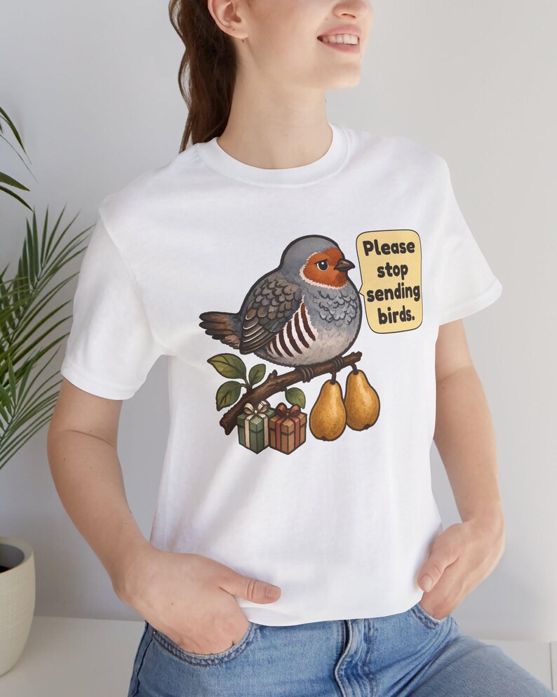 K&ouml;nnte beinhalten: Ein wei&szlig;es T-Shirt mit einer Grafik eines runden Vogels, der auf einem Ast mit zwei Birnen und zwei kleinen Geschenkkartons sitzt. Eine Sprechblase neben dem Vogel enth&auml;lt den Text: Please stop sending birds. Das Design nutzt Erdt&ouml;ne.