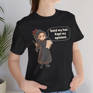 May include: A black short-sleeved t-shirt featuring a cartoon illustration of a person with a long braid, wearing a brown dress and black apron, holding a quill and papers. A speech bubble contains the text: Sold my hair. Kept my opinions.